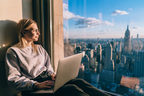 caucasian-female-digital-nomad-sitting-with-laptop-computer-on-window